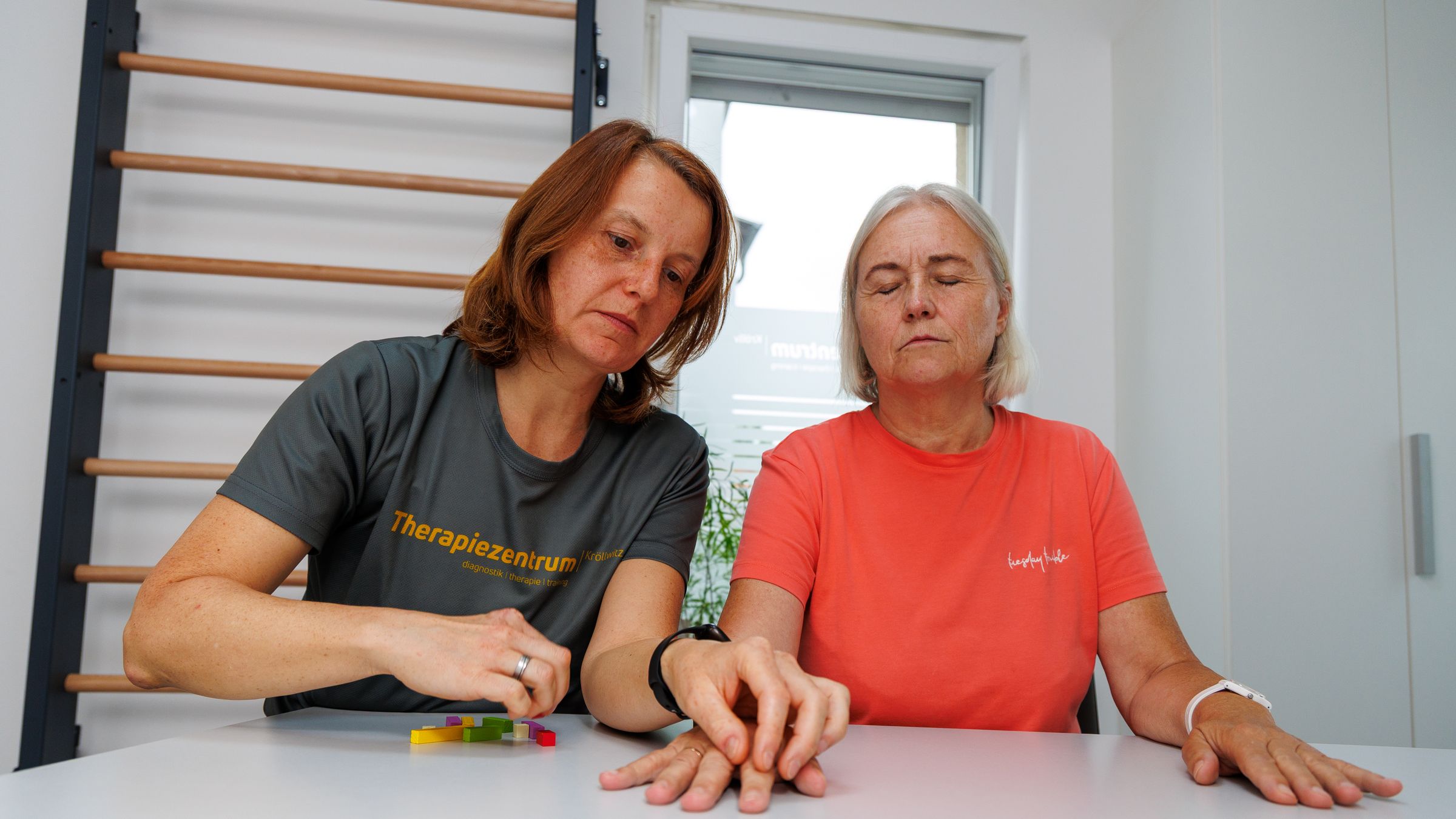 Ergotherapeutin legt einer Patientin am Therapietisch farbige Holzklötze zurecht