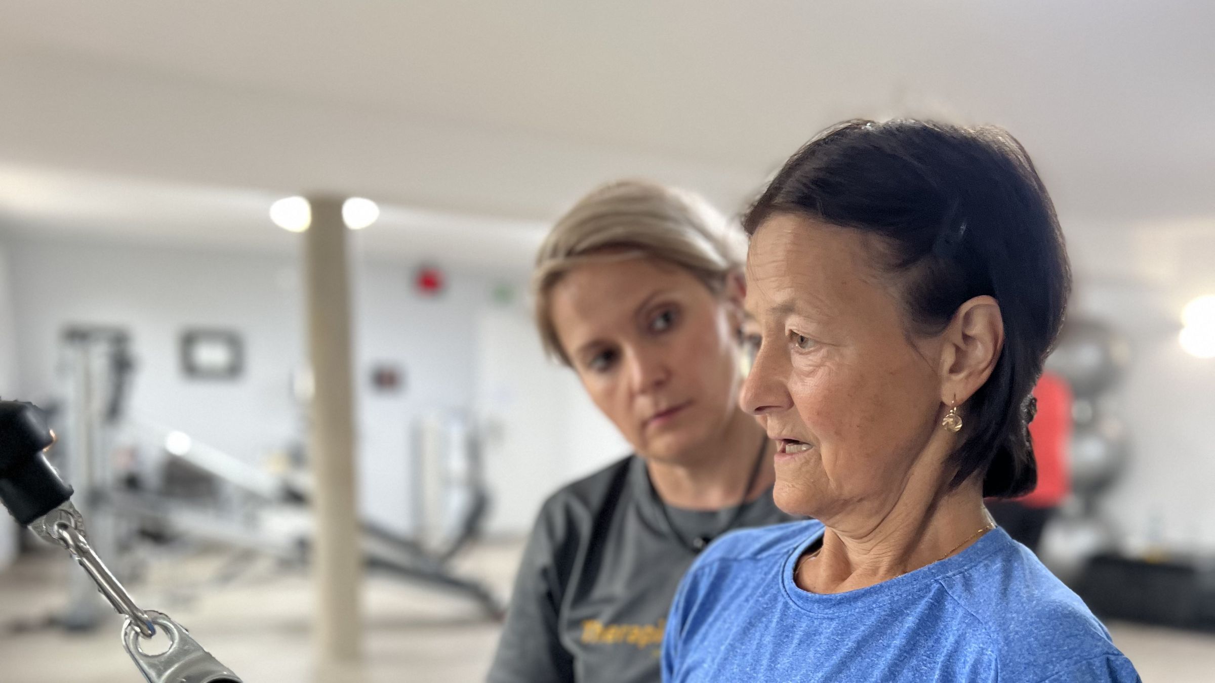 Therapeutin im Praxisshirt korrigiert die Haltung einer Patientin am Seilzuggriff