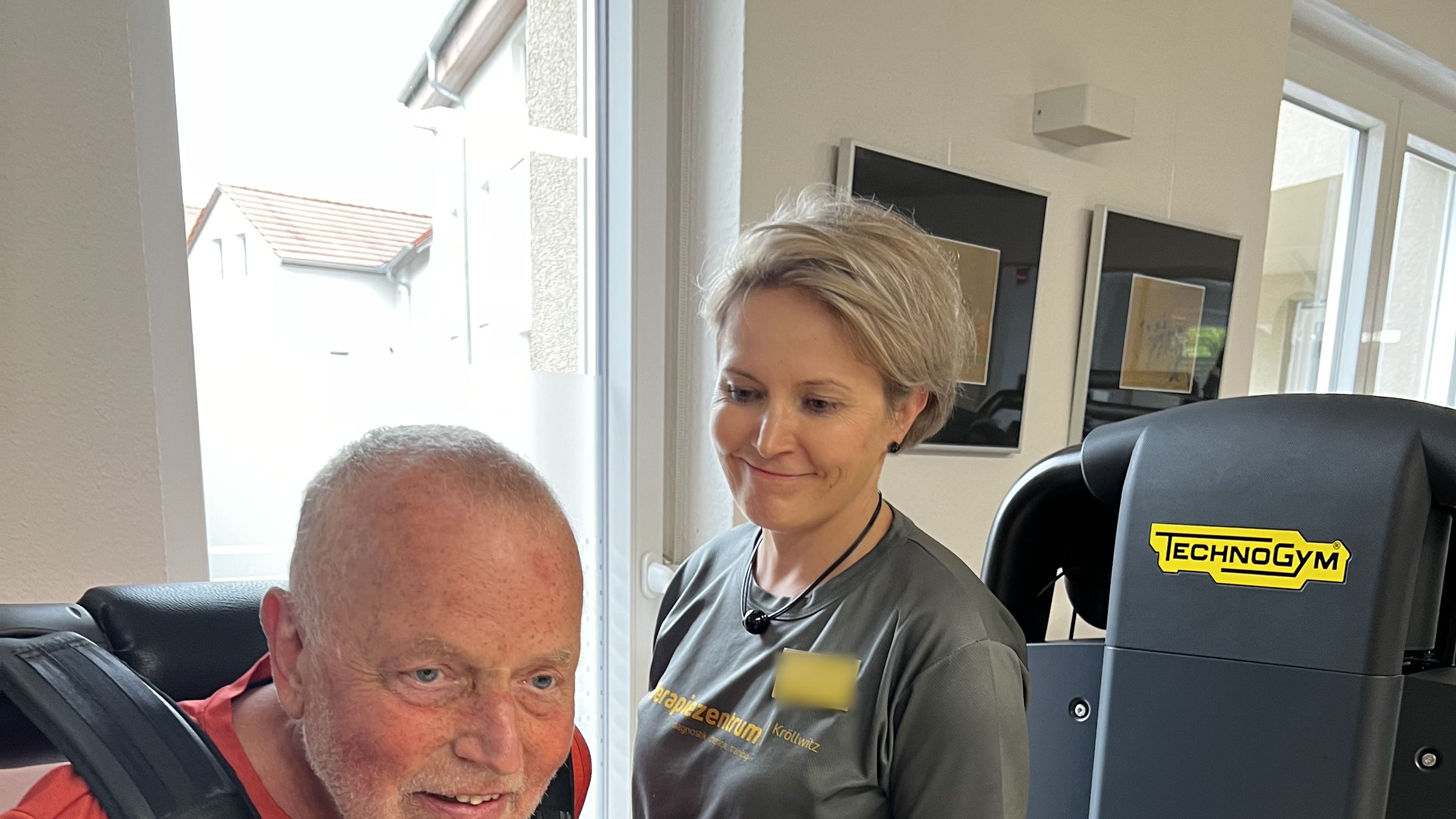 Patient mit Schulterpolstern an einem Technogym-Gerät, die Therapeutin im Praxisshirt steht lächelnd daneben