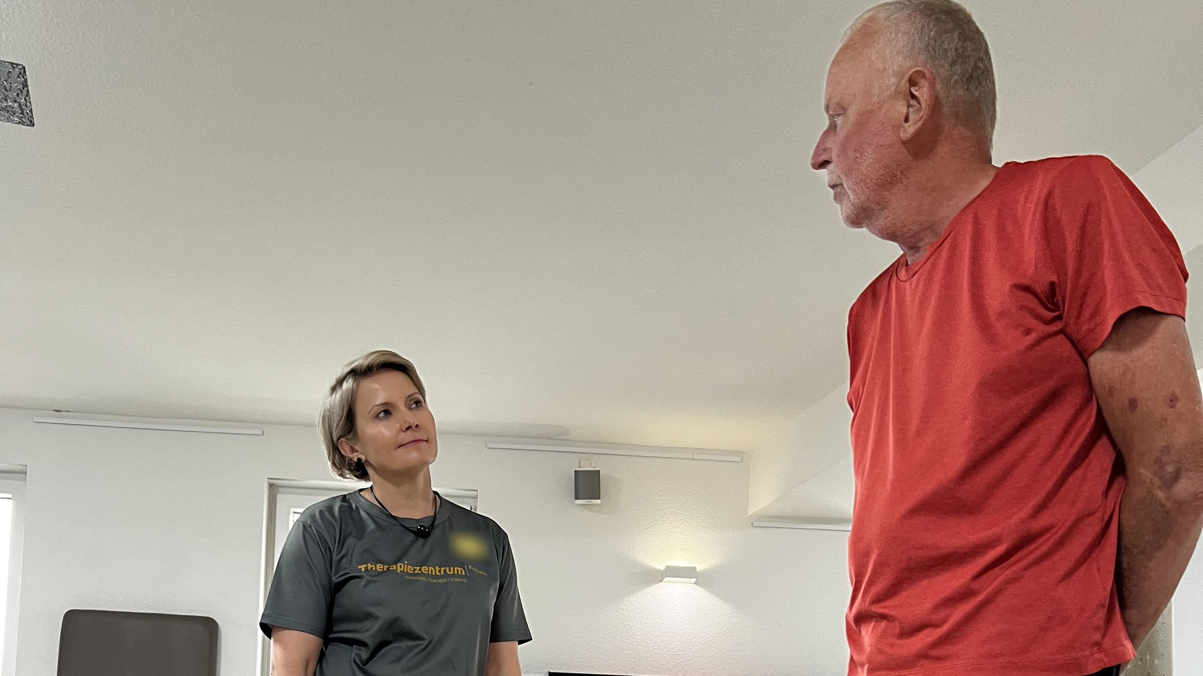 Therapeutin im Praxisshirt im Gespräch mit einem Patienten auf der Trainingsfläche