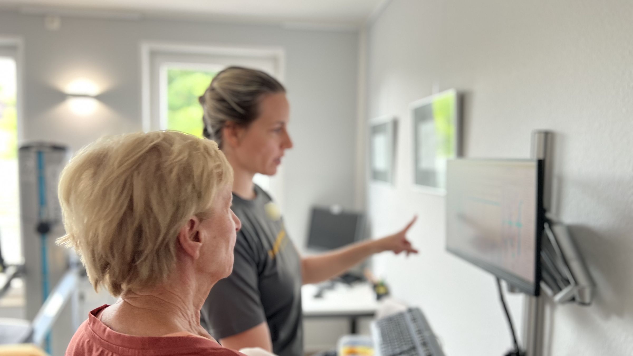 Therapeutin im Praxisshirt zeigt einer Patientin etwas am Bildschirm