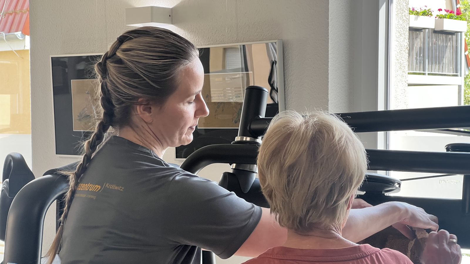Therapeutin im Praxisshirt begleitet eine Patientin bei einer Übung am Trainingsgerät