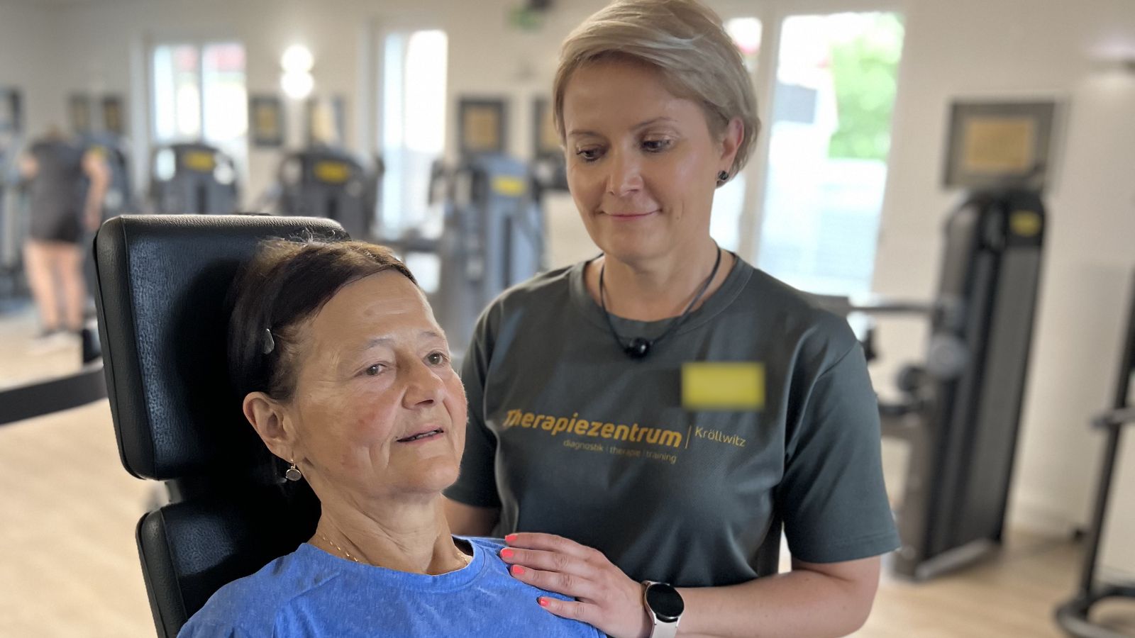 Therapeutin im Praxisshirt legt einer Patientin am Trainingsgerät die Hand auf die Schulter