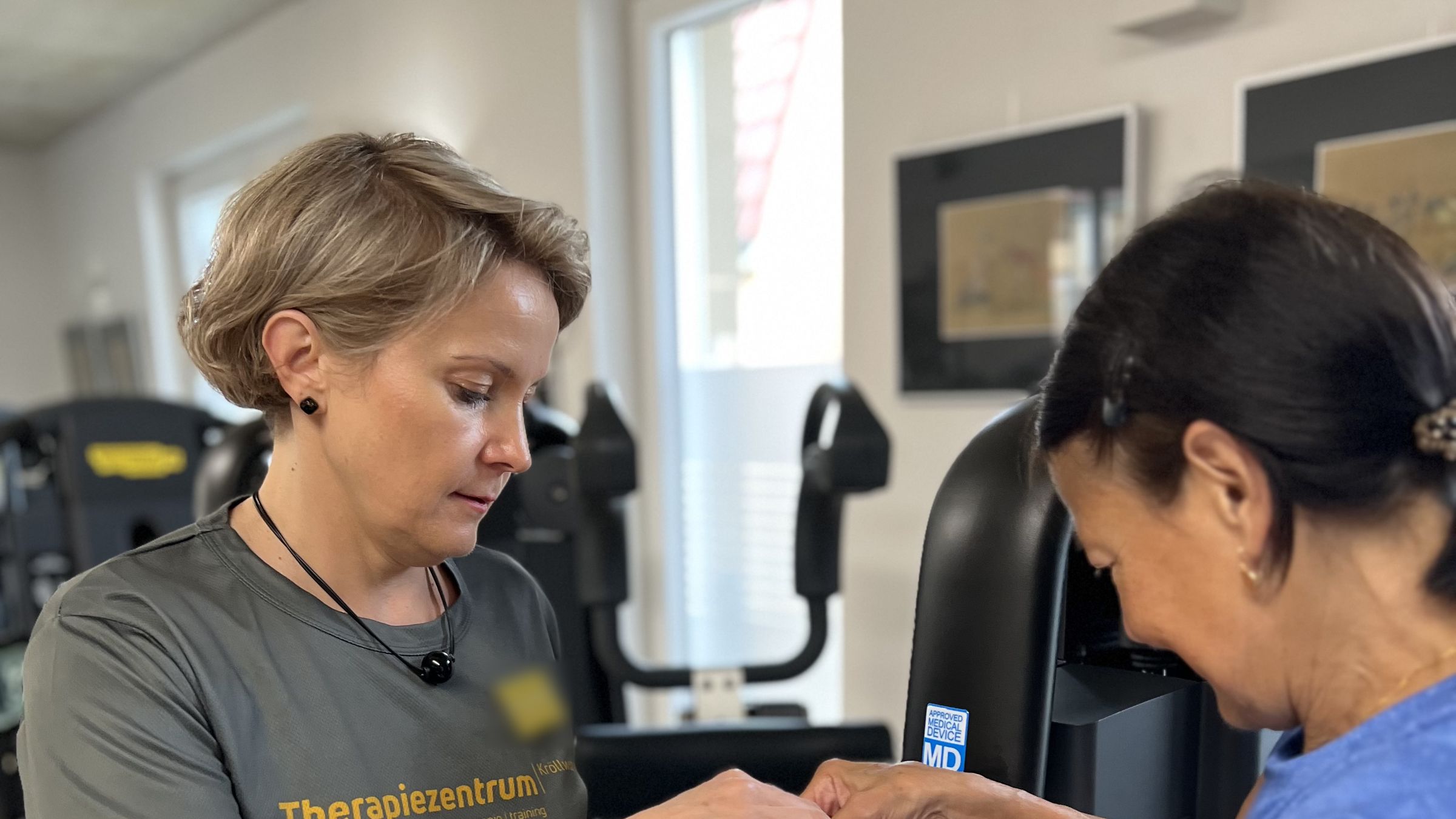Therapeutin im Praxisshirt legt einer Patientin am Trainingsgerät eine Schlaufe um das Handgelenk