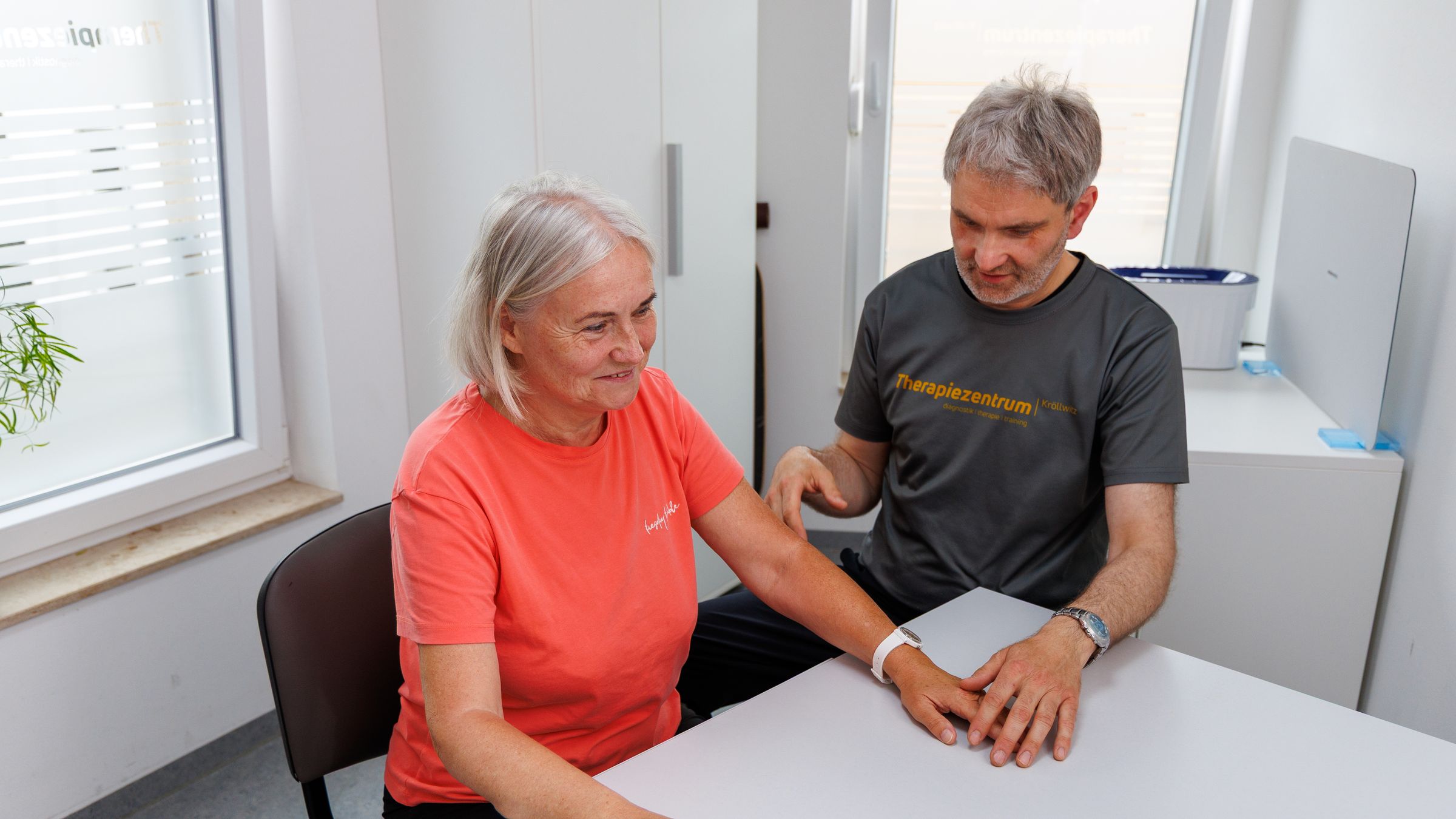 Therapeut im Praxisshirt behandelt am Tisch den Unterarm einer Patientin