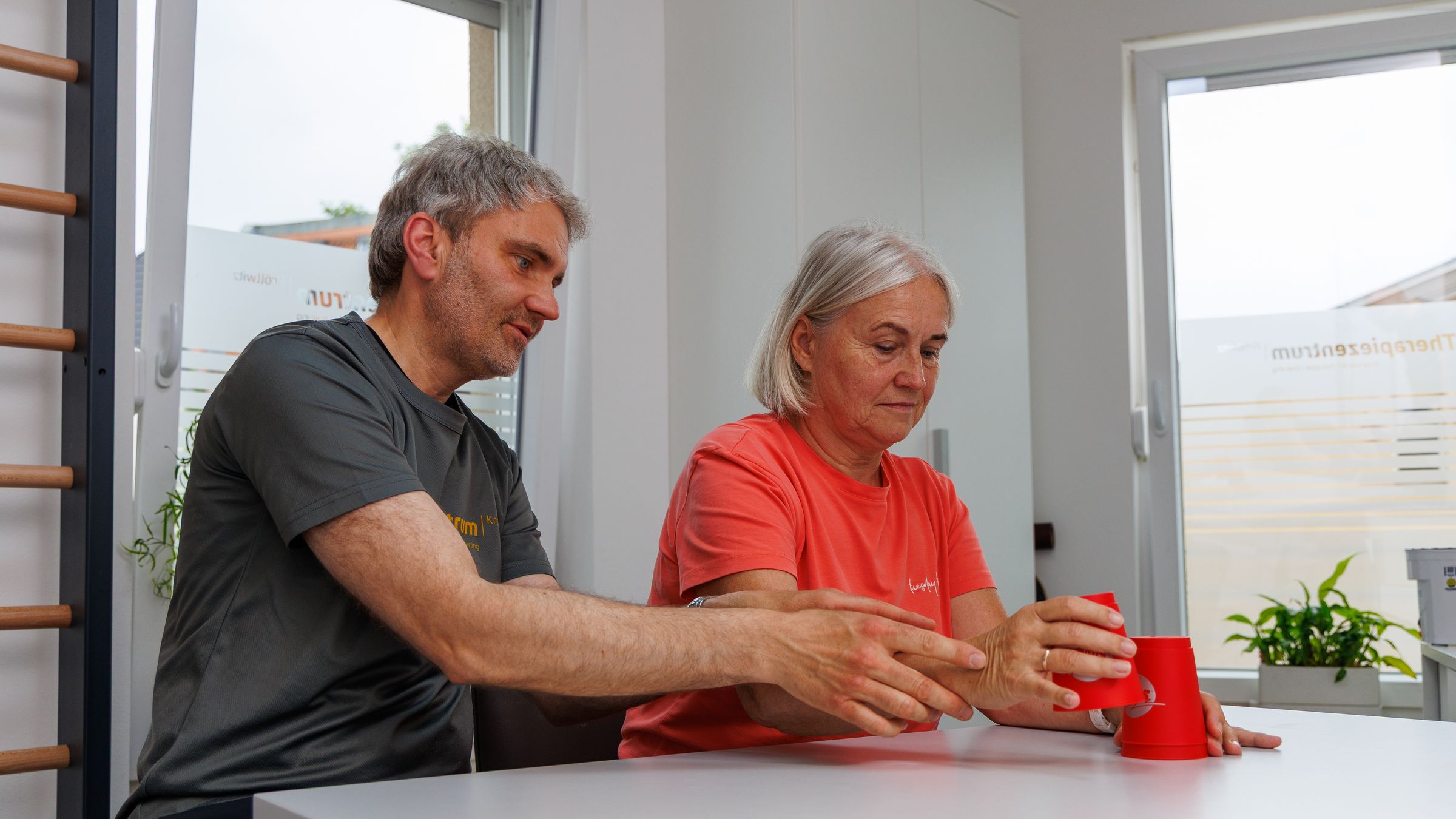 Ergotherapeut führt die Hand einer Patientin beim Greifen eines roten Bechers