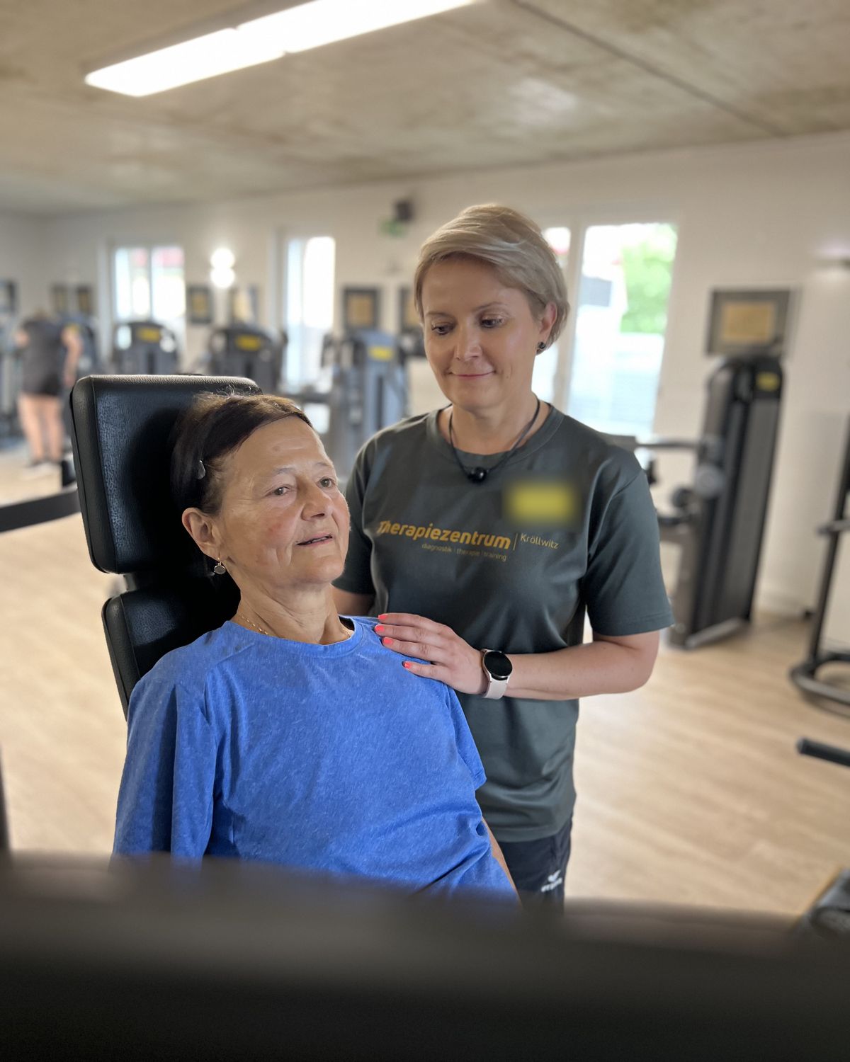 Therapeutin legt einer Patientin im blauen Shirt am Trainingsgerät die Hand auf die Schulter