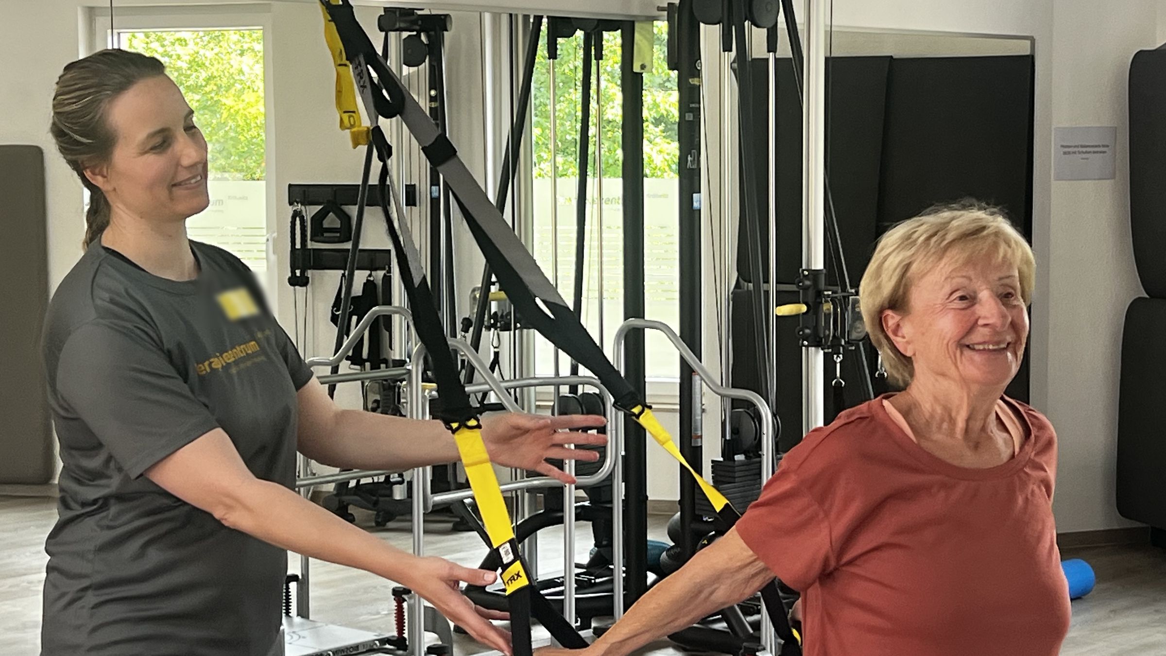 Therapeutin begleitet eine lachende Patientin bei einer Übung am Schlingentrainer auf der Trainingsfläche