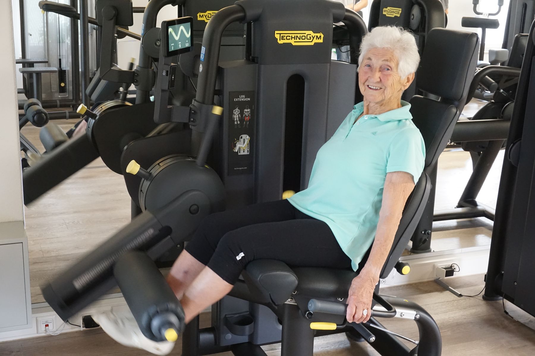 Ältere Patientin trainiert lächelnd an einem Technogym-Beinstrecker